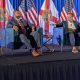 Two men speaking on stage with flags
