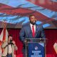 Man speaking at podium with American flag backdrop