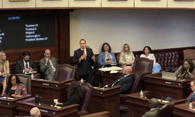 Lawmakers debate in legislative chamber with vote board