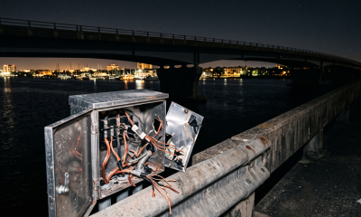 A close-up view of an opened electrical utility box on a concrete bridge railing at night, showing cut and exposed copper wires from a theft incident. In the background is the dark Barrancas Bridge over Bayou Chico with distant city lights.