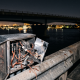 A close-up view of an opened electrical utility box on a concrete bridge railing at night, showing cut and exposed copper wires from a theft incident. In the background is the dark Barrancas Bridge over Bayou Chico with distant city lights.