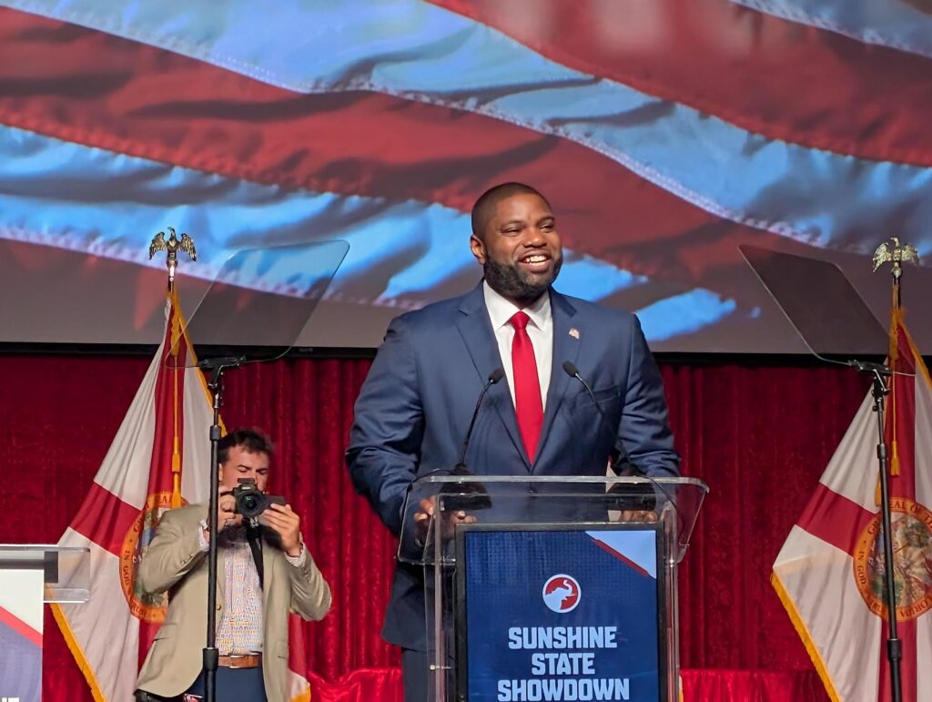 Man speaking at podium with American flag backdrop