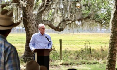 Man speaking at outdoor podium under oak trees