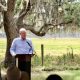 Man speaking at outdoor podium under oak trees