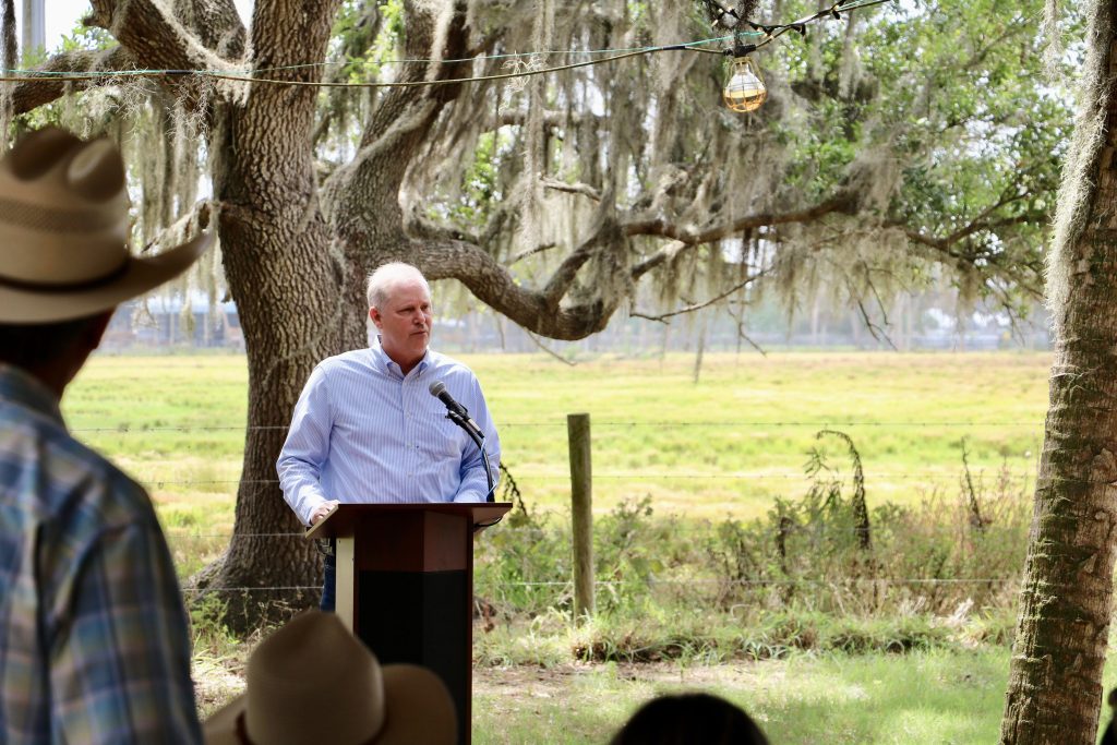 Man speaking at outdoor podium under oak trees