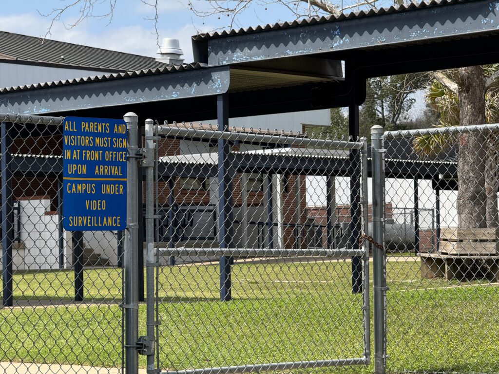 School campus behind chain-link fence with surveillance sign