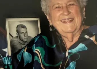 Elderly woman smiling beside framed black-and-white portrait