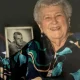 Elderly woman smiling beside framed black-and-white portrait