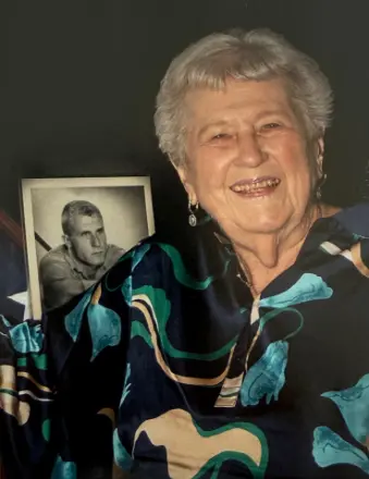 Elderly woman smiling beside framed black-and-white portrait