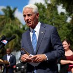 Charlie Crist speaking at a podium with microphones, flanked by supporters and palm trees — Charlie Crist mayoral campaign