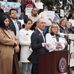 Democratic lawmakers speaking at a press conference with supporters holding signs.