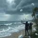 Two figures in raincoats point towards a stormy sea with lightning, while ice pellets fal — alerta meteorologica pensacola