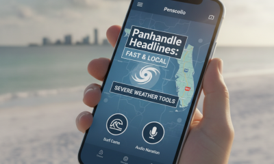 A hand holding a smartphone displaying the Newswk App Pensacola interface with weather tools and headlines against a beach…