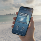 A hand holding a smartphone displaying the Newswk App Pensacola interface with weather tools and headlines against a beach…