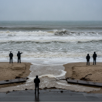 Five individuals in dark jackets observing rough ocean waves on a beach, highlighting rip cur — rip current risk Pensacola