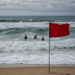 Red warning flag on a beach as swimmers face rough surf during a rip current state — rip current statement escambia county