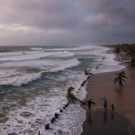 People standing near a beach with rough waves and palm trees, highlighting the Pensacola rip current warning.