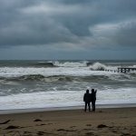 Two people standing on a beach watching large waves under a cloudy sky, indicating a Pensacola Beach high surf warning.