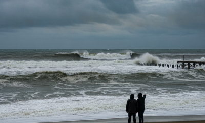 Two people standing on a beach watching large waves under a cloudy sky, indicating a Pensacola Beach high surf warning.